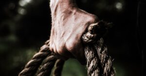 A close-up image shows a strong, firm hand gripping a thick, rough rope, showcasing strength and determination in an outdoor setting.