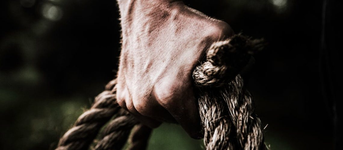 A close-up image shows a strong, firm hand gripping a thick, rough rope, showcasing strength and determination in an outdoor setting.